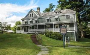 Historic gray house with a large porch, steps leading up a grassy hill, and a historical marker sign.